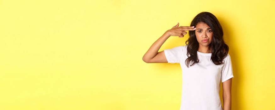 Portrait of skeptical and bothered african-american woman, making finger gun sign over head and smirking unamused, standing over yellow background.