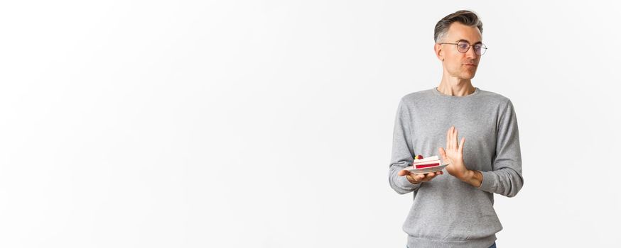 Portrait of displeased middle-aged man, looking away disgusted and rejecting piece of cake, standing bothered over white background.