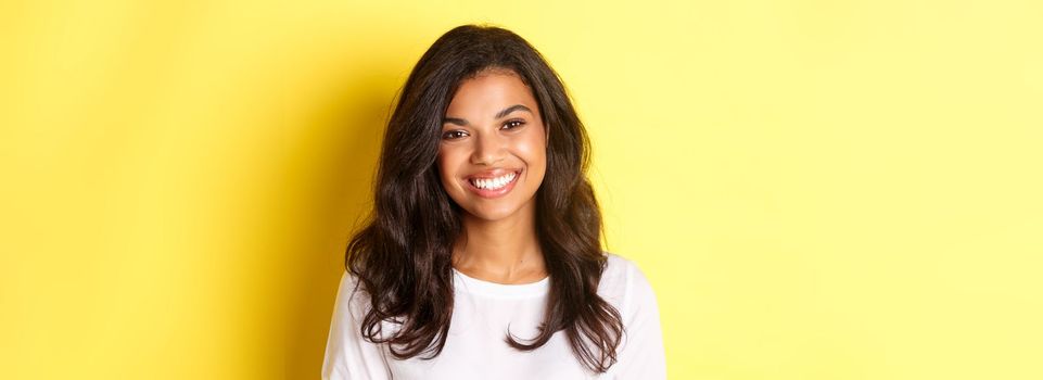 Close-up of attractive african-american woman, smiling and looking happy, standing over yellow background.