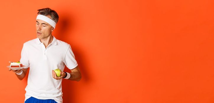 Portrait of middle-aged sportsman making decision between green apple and cake, being on diet, standing over orange background.