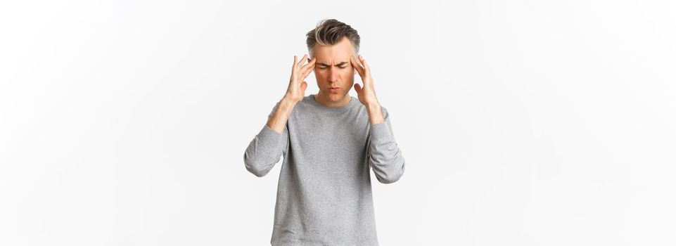 Portrait of middle-aged man in grey sweater having a headache, touching head and grimacing from painful migraine, standing over white background.