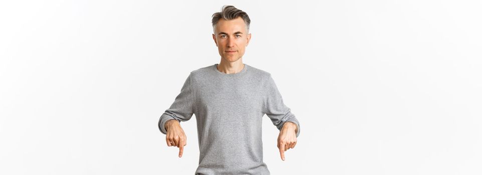 Portrait of successful middle-aged man in grey sweater, pointing fingers down and looking confident at camera, showing logo, standing over white background.