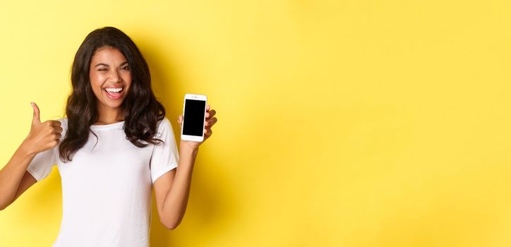 Image of beautiful african-american girl, winking and smiling, showing smartphone screen and thumbs-up in approval, standing over yellow background.