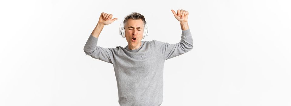 Portrait of handsome, modern middle-aged man, listening music in headphones and dancing, standing over white background.