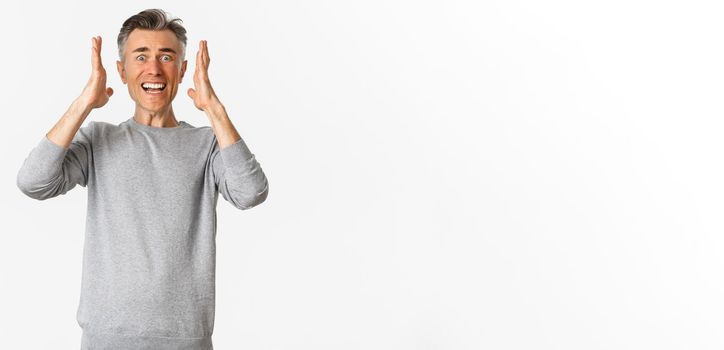 Image of frustrated middle-aged guy in grey sweater panicking, shaking hands and looking distressed, standing over white background.