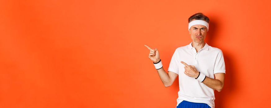 Portrait of disappointed middle-aged man in sportswear, complaining and pointing fingers left at logo, showing something bad, standing over orange background.
