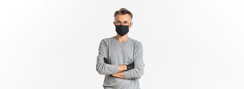 Concept of coronavirus, lifestyle and quarantine. Portrait of displeased middle-aged man in medical mask and gloves, cross arms on chest and looking disappointed, standing over white background.
