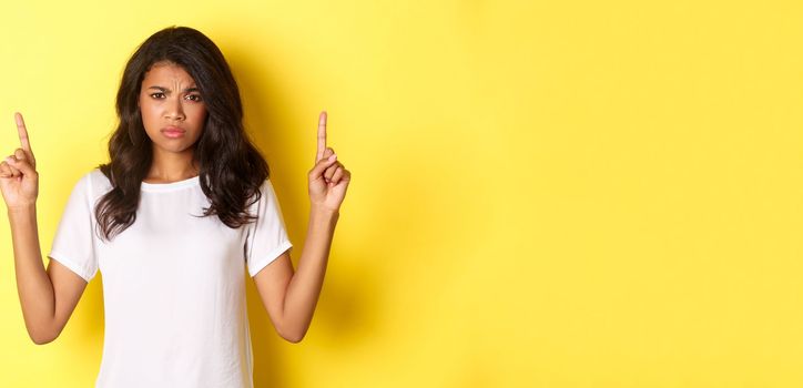 Portrait of upset and disappointed teenage african-american girl, complaining and frowning, pointing fingers up at something upsetting, standing over yellow background.