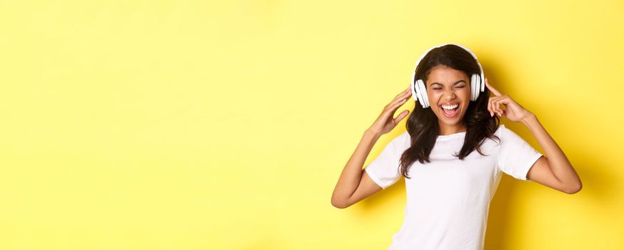 Portrait of modern african-american girl, enjoying listening to music in headphones, smiling amazed, standing over yellow background.