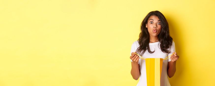 Image of surprised african-american girl receive a gift on holiday, open a shopping bag and looking amazed, standing over yellow background.