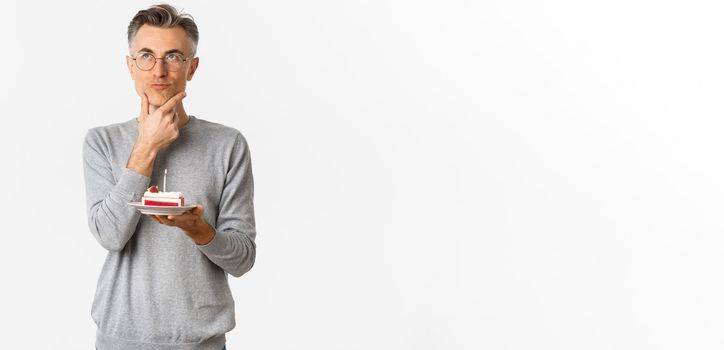 Image of thoughtful and serious middle-aged man, celebrating birthday, holding b-day cake and thinking what wish to make, standing over white background.