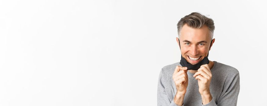 Close-up of handsome middle-aged man, taking-off medical mask and looking happy, smiling pleased, standing over white background. Concept of covid-19, social distancing and quarantine.