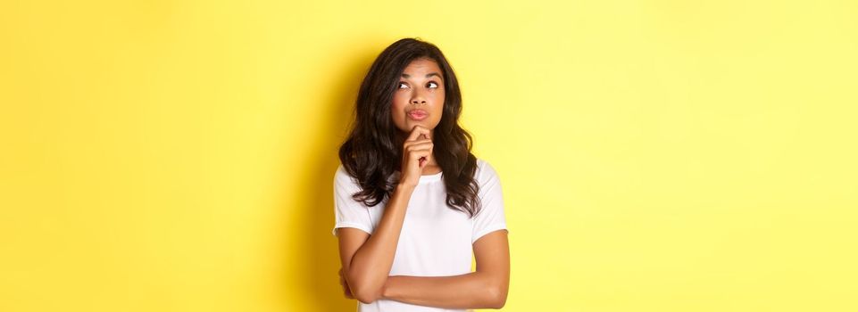 Image of thoughtful, beautiful african american girl making her choice, looking at upper right corner and thinking, standing over yellow background.