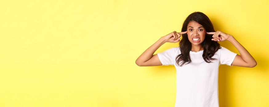 Portrait of annoyed and angry girl in white t-shirt, complaining on loud noise, shut ears and looking mad, standing over yellow background.