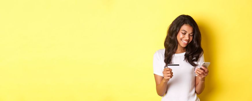 Portrait of good-looking african american woman, shopping online with smartphone and credit card, standing over yellow background.