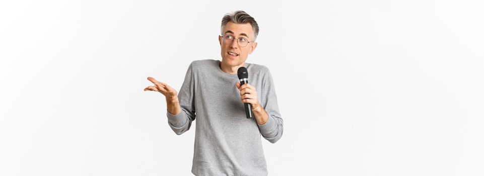 Portrait of handsome middle-aged man in glasses and gray sweater, making speech, talking with microphone and explaining something, standing over white background.