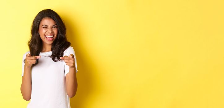 Portrait of cheeky, beautiful african-american woman in white t-shirt, winking and pointing fingers at camera to say congrats, praising you, standing over yellow background.