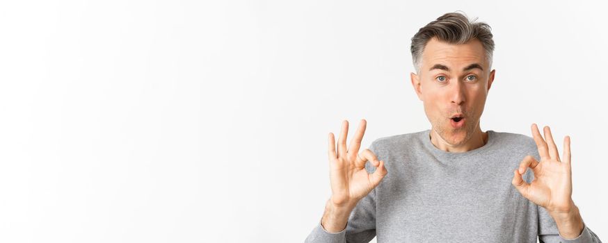 Close-up of impressed middle-aged guy, showing okay signs in approval, like something good, recommending product, standing over white background.