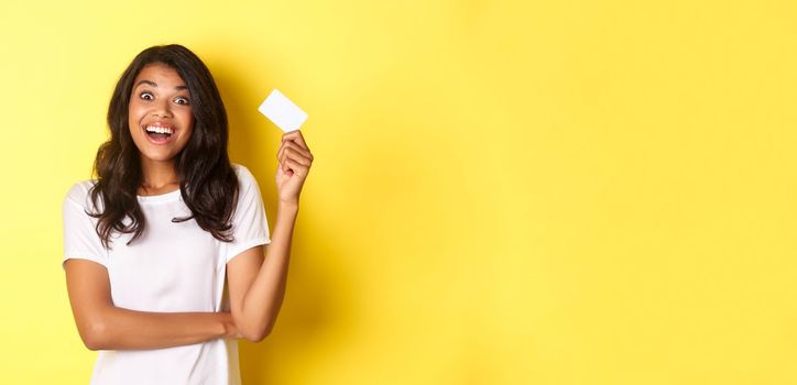 Portrait of amazed african-american girl, raising credit card and smiling excited, going on shopping, standing over yellow background.