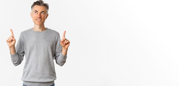 Image of handsome middle-aged man in grey sweater making his choice, smiling thoughtful, pointing fingers up and looking at product, standing over white background.