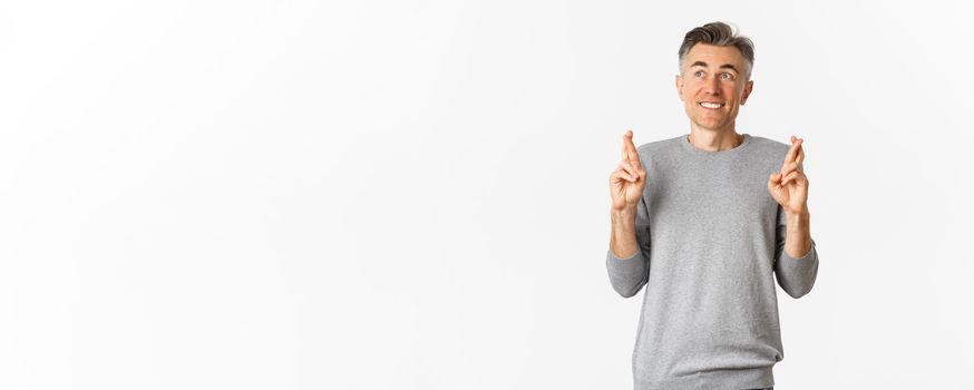 Image of excited and hopeful middle-aged man making a wish, waiting for results, cross fingers for good luck and looking at upper left corner, standing over white background.