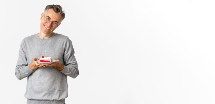 Portrait of smiling middle-aged man, looking touched and happy, celebrating birthday, holding b-day cake and celebrating, standing over white background.