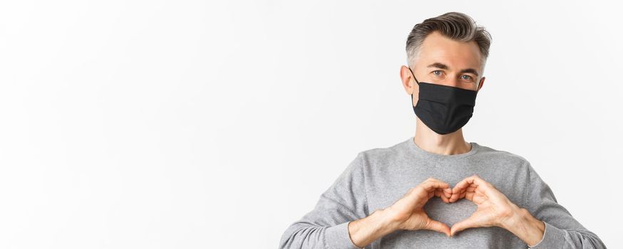 Concept of covid-19, social distancing and quarantine. Close-up of attractive middle-aged man in black medical mask, showing heart sign and smiling, standing over white background.