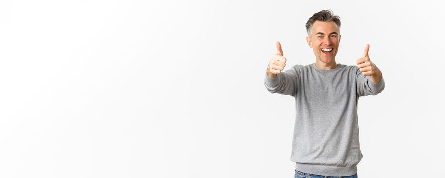Portrait of cheerful middle-aged man praising good work, standing over white background, showing thumbs-up in approval and smiling glad.
