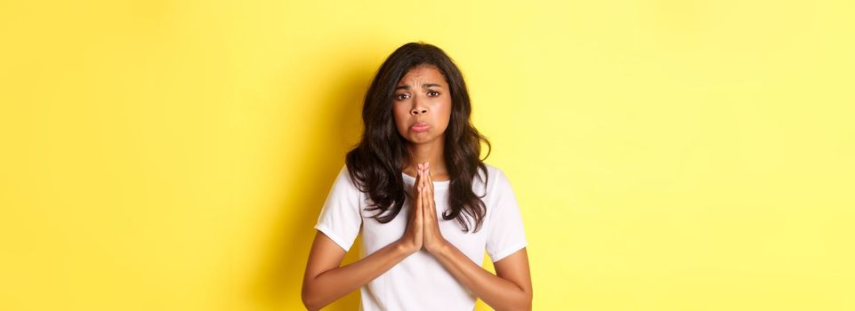 Portrait of sad african-american girl pleading, begging for help and sulking upset, standing over yellow background.