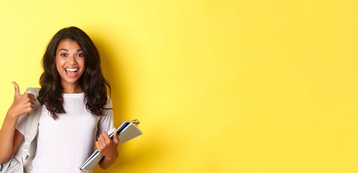 Image of cheerful african-american girl student, holding notebooks for classes and backpack, showing thumbs-up, like studying, standing over yellow background.