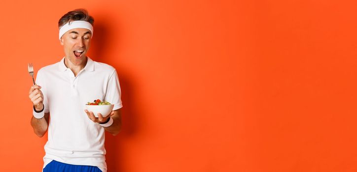 Concept of sport, fitness and lifestyle. Portrait of joyful middle-aged guy in workout uniform, holding fork and salad, eating healthy food, standing over orange background.