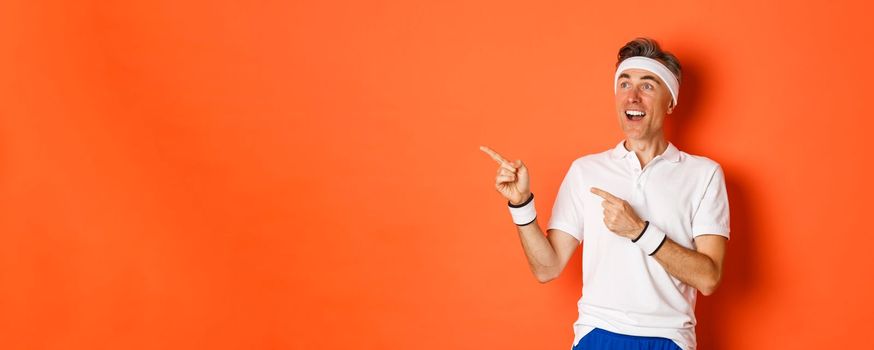 Portrait of amazed middle-aged male athlete, pointing fingers and looking left at gym promo, working out over orange background.