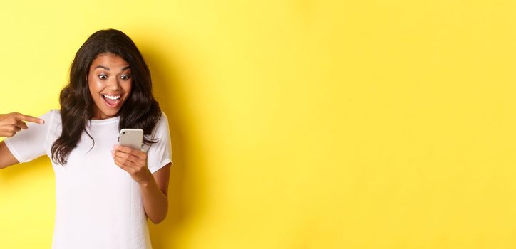 Image of excited african-american girl, looking and pointing finger at mobile phone, seeing something amazing online, standing over yellow background.