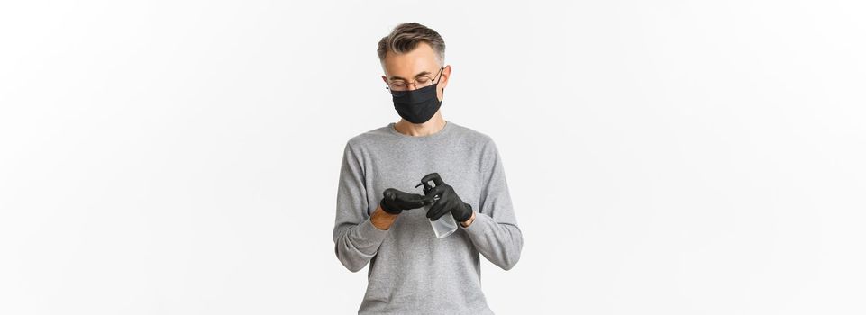 Concept of coronavirus, lifestyle and quarantine. Image of middle-aged man in medical mask and gloves, applying hand sanitizer, preventing catching covid-19, white background.