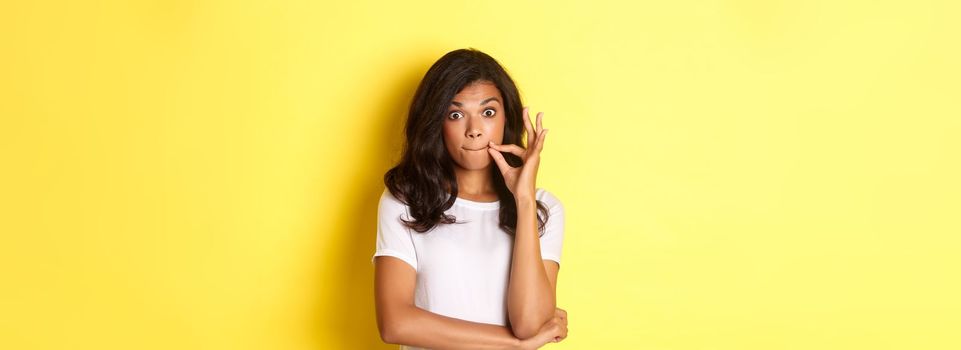 Portrait of young african-american woman making promise to keep secret, seal lips, zipping mouth with fingers, standing over yellow background.