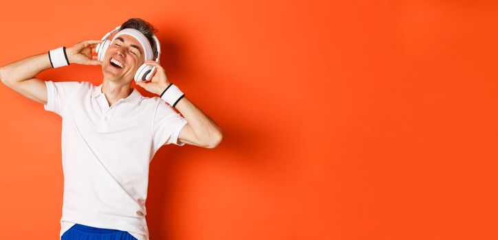 Portrait of carefree middle-aged male athlete, listening music in headphones during fitness training, standing over orange background.