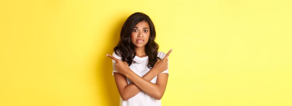 Portrait of indecisive african-american girl need help with choice, pointing fingers sideways and biting lip in doubt, asking advice, standing over yellow background.