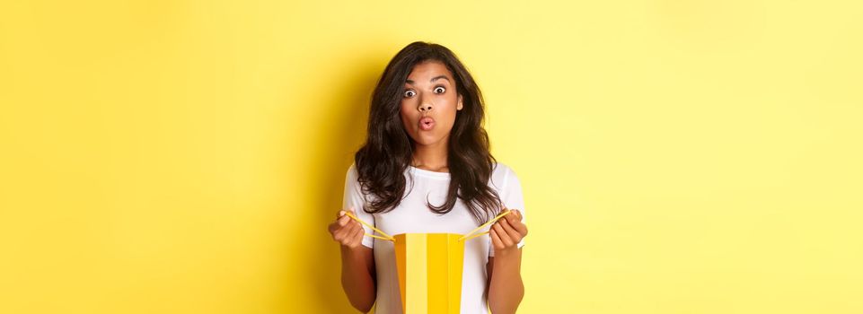 Image of surprised african-american girl receive a gift on holiday, open a shopping bag and looking amazed, standing over yellow background.