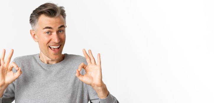 Close-up of handsome middle-aged man, showing okay signs and looking amazed, recommending something cool, standing over white background.