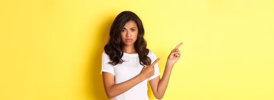 Portrait of displeased and worried african-american woman, expressing her doubts, pointing fingers at upper left corner and frowning hesitant, standing over yellow background.