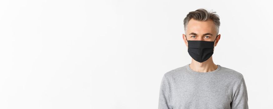 Concept of covid-19, social distancing and lifestyle. Close-up of handsome middle-aged man in black medical mask, looking confident at camera, standing over white background.