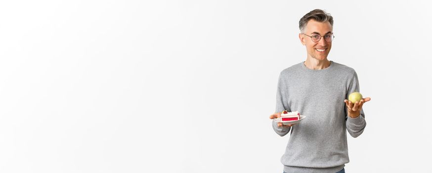 Image of handsome and healthy middle-aged man in glasses, recommending eat apple instead of cake, standing over white background.