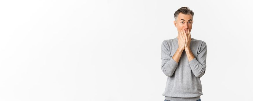 Image of excited and amused caucasian man with gray hair, cover mouth and looking at something interesting, standing over white background.