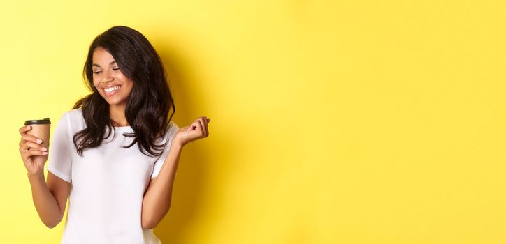 Portrait of happy and pleased african-american girl, looking at coffee and smiling, standing over yellow background.