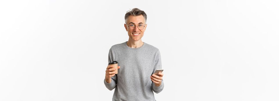 Handsome middle-aged man in grey sweater and glasses, smiling happy, drinking coffee and using mobile phone, standing over white background.