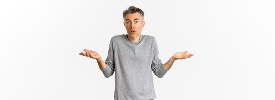 Image of confused middle-aged man shrugging shoulders, looking clueless at camera, standing over white background.