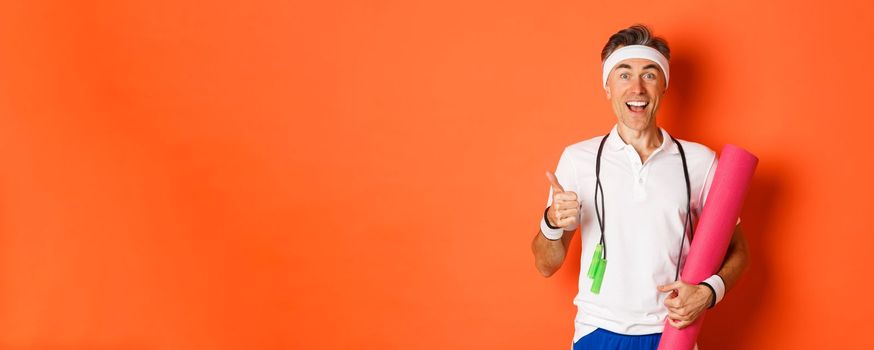 Concept of workout, gym and lifestyle. Cheerful middle-aged fitness guy, holding skipping rope and yoga mat, showing thumbs-up and smiling satisfied, standing over orange background.