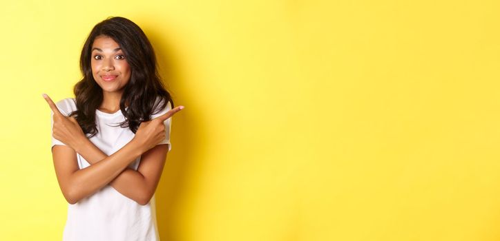 Portrait of cute indecisive african american girl, pointing fingers sideways and shrugging, asking advice with choices, standing over yellow background.