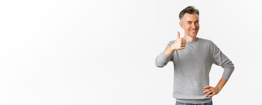 Portrait of proud and happy middle-aged man, smiling pleased and showing thumbs-up, praise something good, standing over white background.