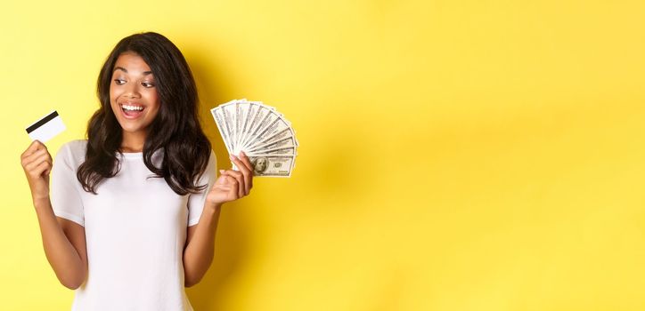 Portrait of attractive african-american girl, holding money and looking at credit card, standing over yellow background.